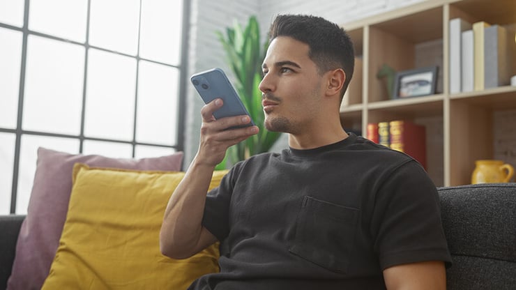 Man using a smartphone to interact with an IVR system, speaking into the device while sitting on a couch in a modern living room.