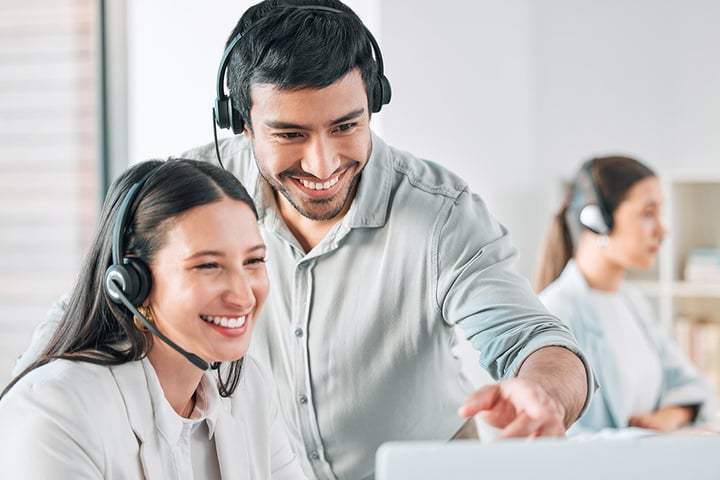 Smiling call centre agents working together, wearing headsets, with a colleague in the background